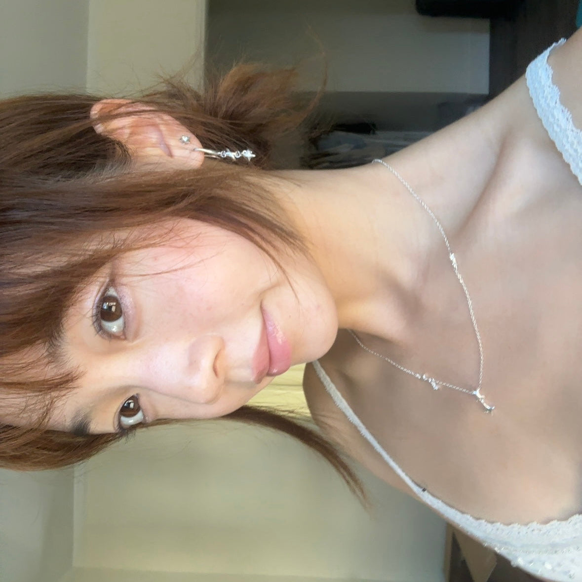 A close-up selfie of a woman wearing a delicate silver necklace and matching silver stud earrings in a domestic indoor setting.