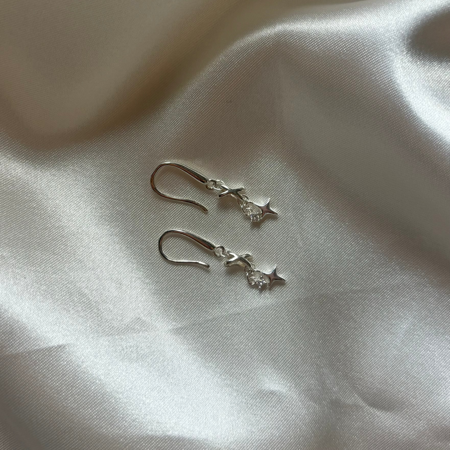 A pair of silver dangle earrings with a small star charm, resting on a draped white satin fabric background.