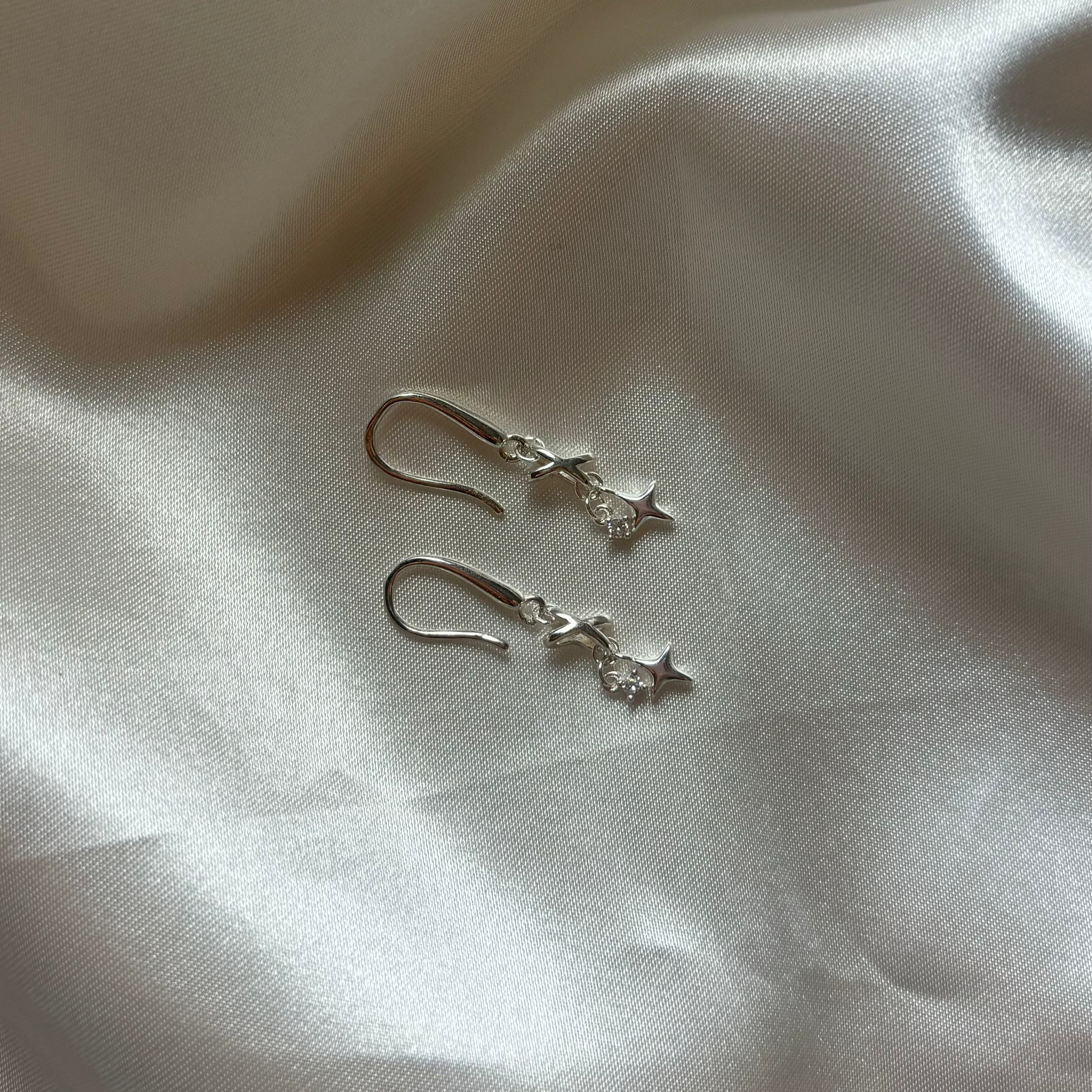 A pair of silver dangle earrings with a small star charm, resting on a draped white satin fabric background.