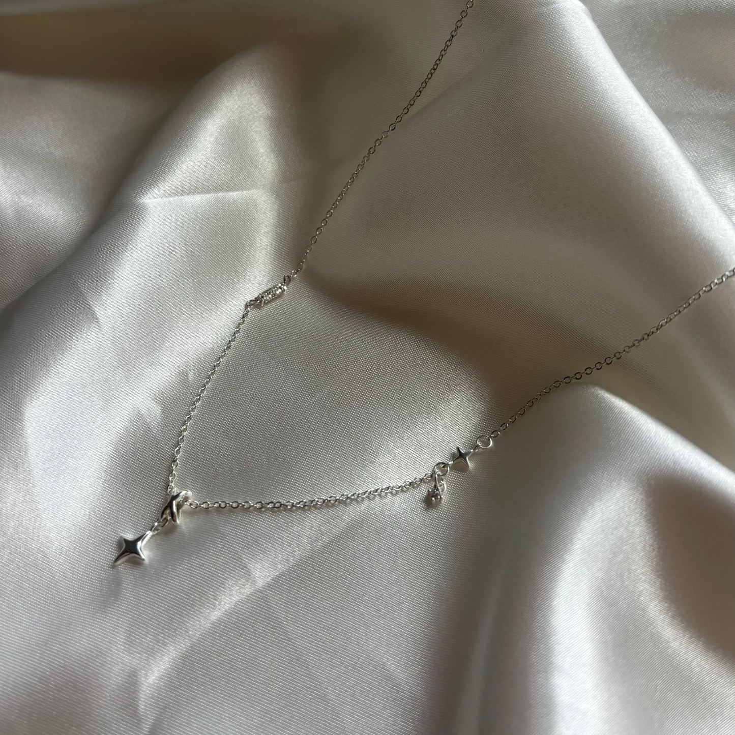 A delicate silver necklace with three small star and crescent moon charms, displayed on draped white satin fabric.