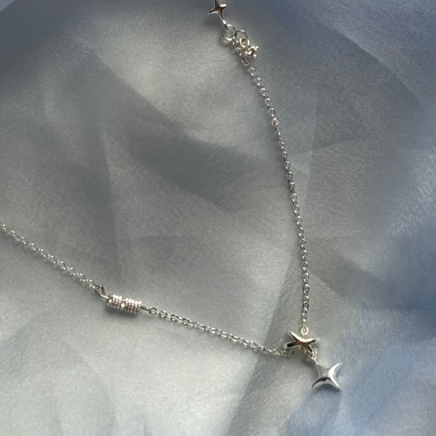 A delicate silver necklace with small star and bead details, draped over light blue satin fabric.