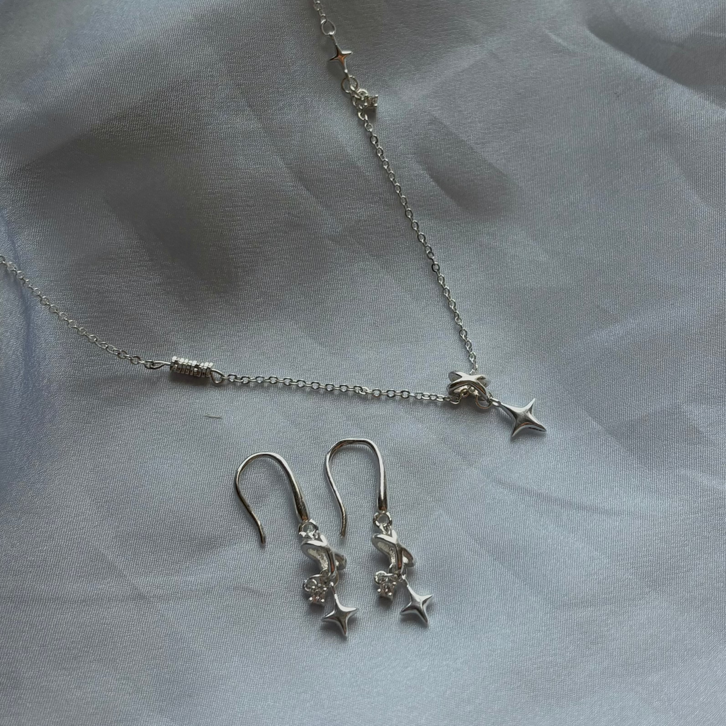 A silver jewelry set featuring a delicate chain necklace and matching drop earrings, both adorned with small star charms, displayed on a soft, draped white fabric background.