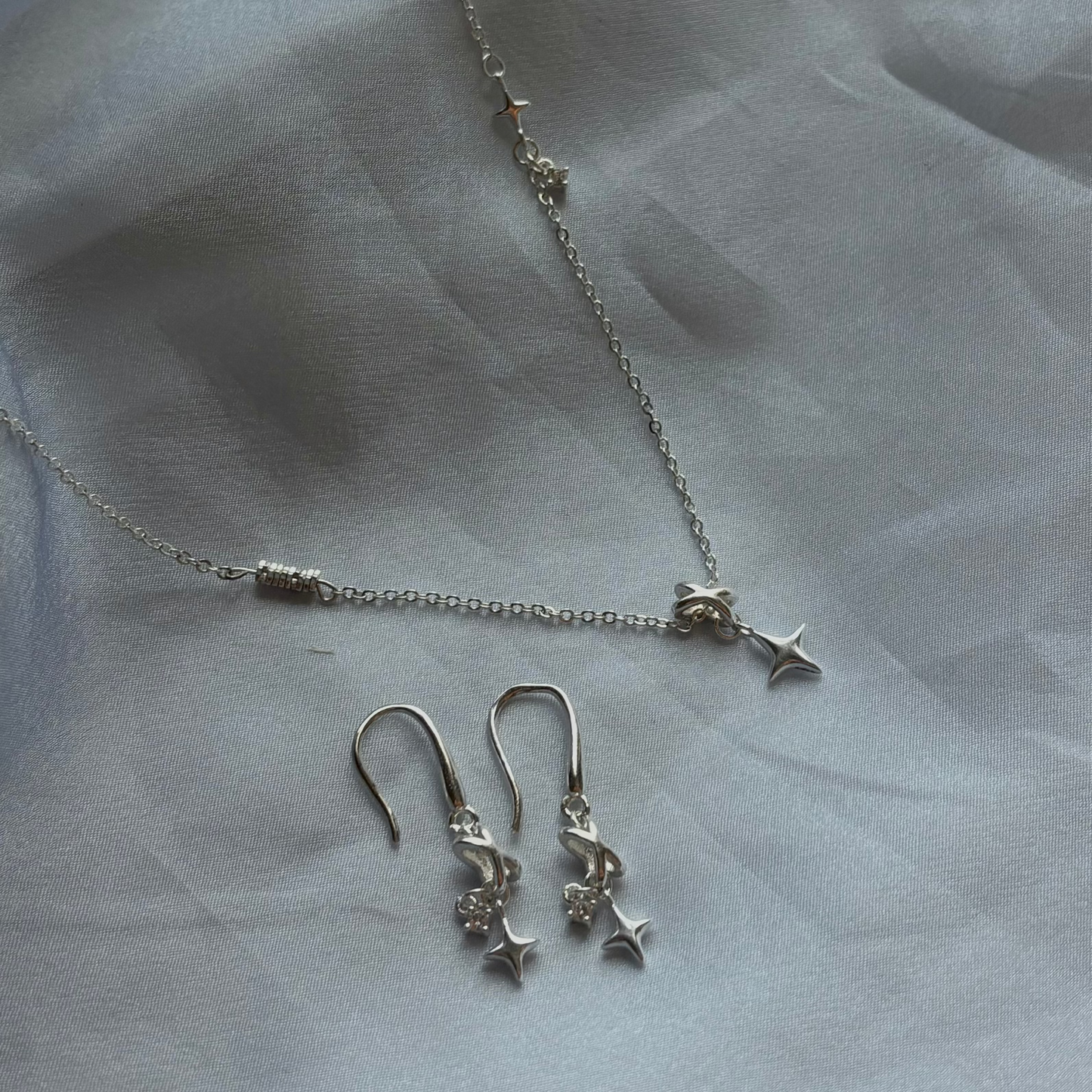 A silver jewelry set featuring a delicate chain necklace and matching drop earrings, both adorned with small star charms, displayed on a soft, draped white fabric background.