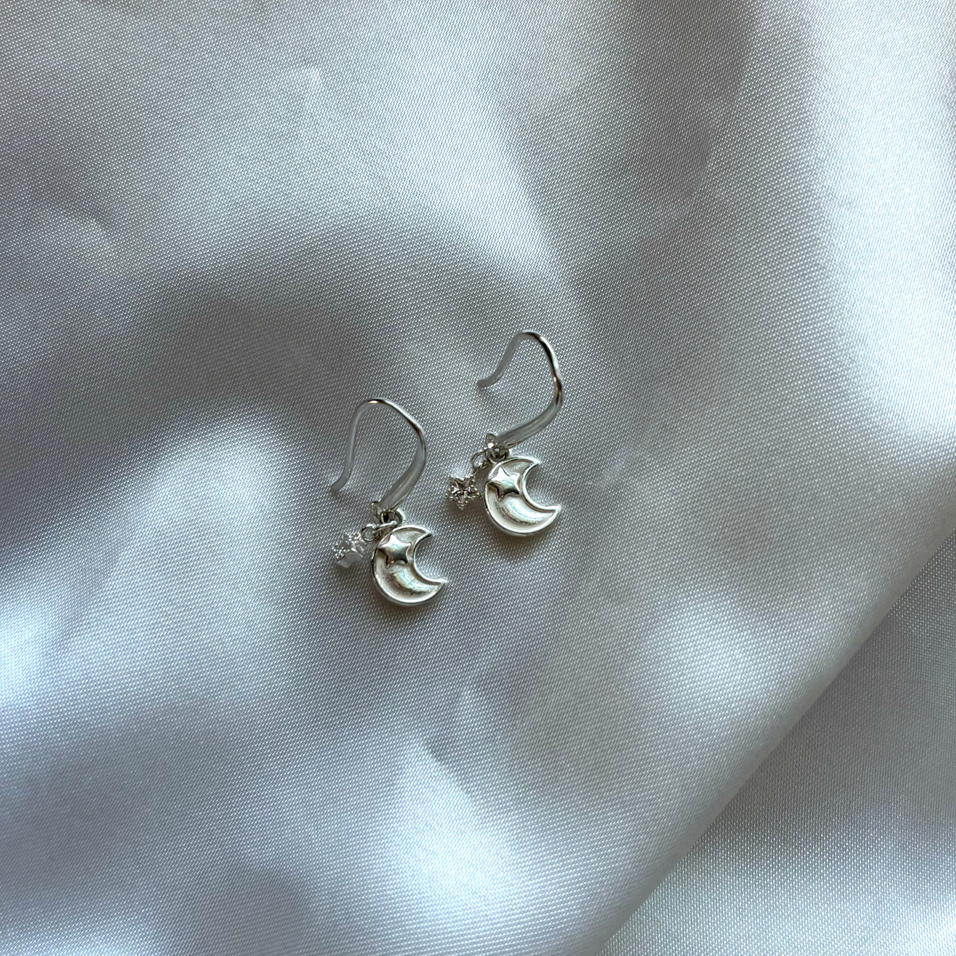 A pair of silver crescent moon and star dangle earrings resting on draped white satin fabric.
