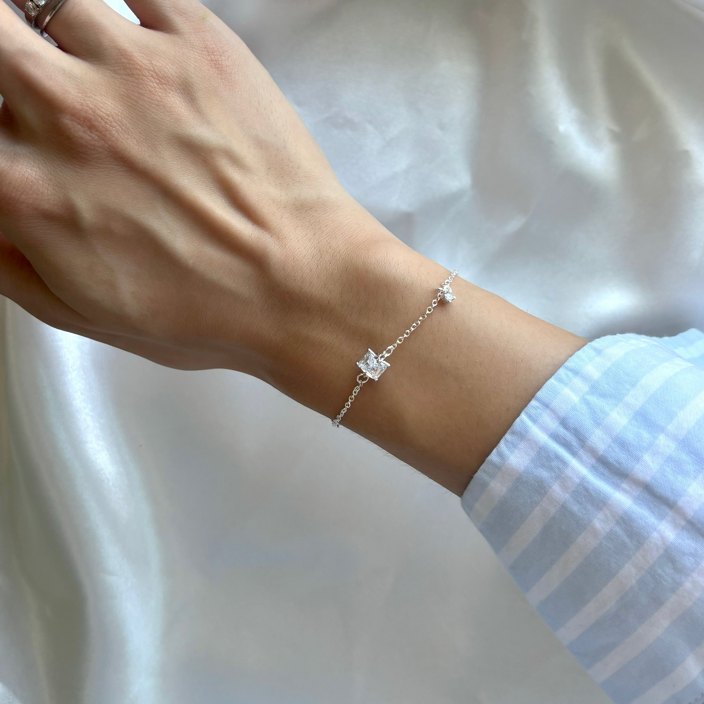 A delicate silver chain bracelet with a small clear crystal charm worn on a person's wrist, shown against a draped white satin fabric background.