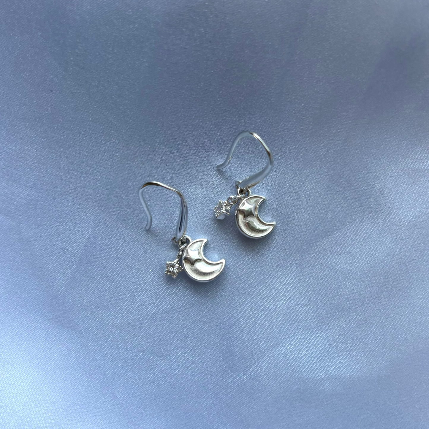 A pair of silver crescent moon and star dangle earrings resting on a soft, light blue satin fabric background.