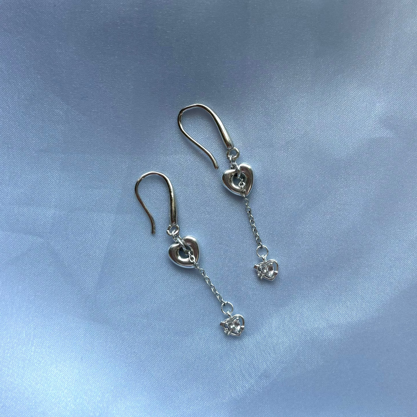 A pair of silver dangle earrings with heart-shaped charms and small clear crystal drops, displayed on a soft, light blue satin fabric background.