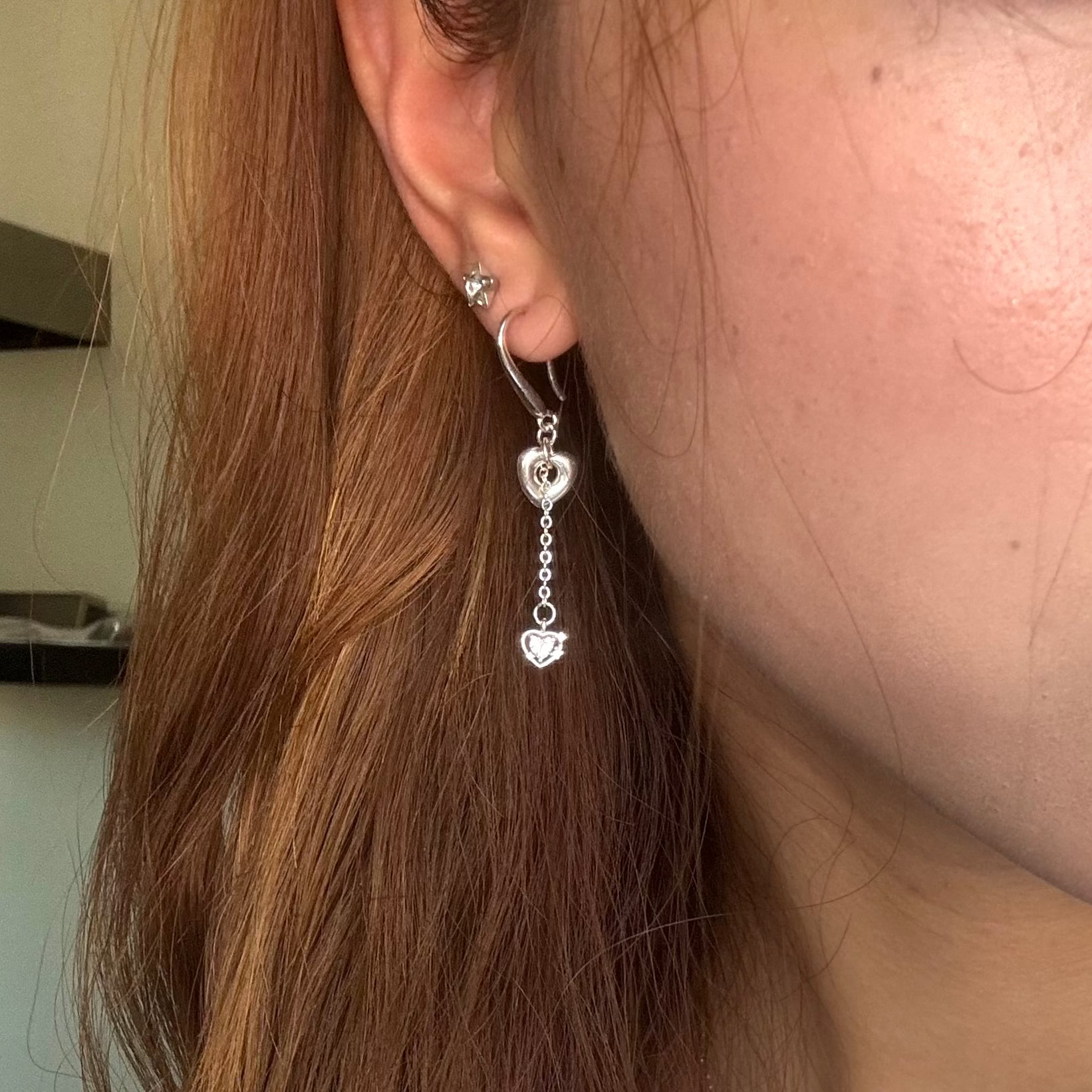 A close-up of a person's ear wearing a silver heart earring with a dangling heart-shaped charm, shown in a casual indoor setting.