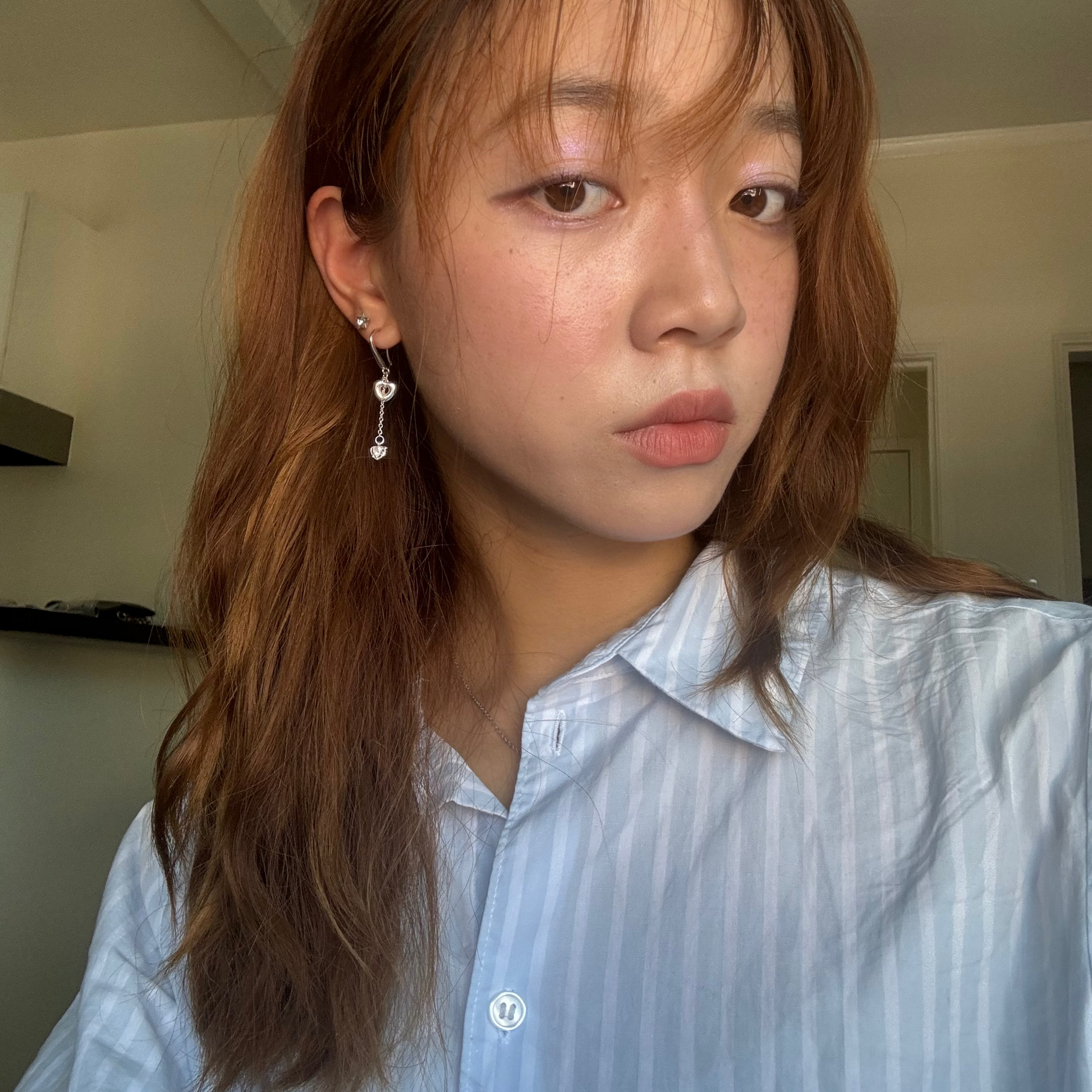 A close-up selfie of a woman with light brown hair wearing a light blue striped button-up shirt and a silver heart dangle earring with a small sparkly heart detail.