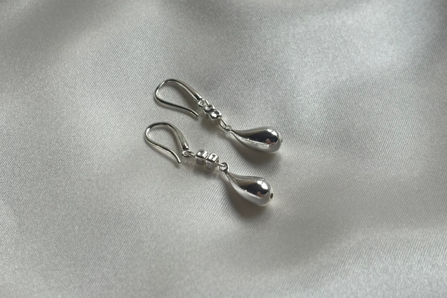 A pair of handmade sterling silver teardrop-shaped earrings laid on a silky surface.